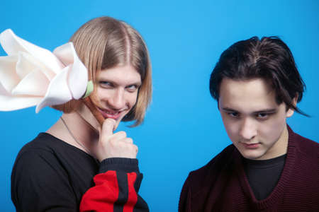 Two young handsome men with funny face expressions, friends or couple. Gay relationship, diverse family. One with ginger blond hair, smiling squinty, the other is serious. Blue background, soft focusの写真素材
