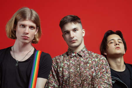 Three young handsome men of different nationalities stand on vibrant red background. Gay relationship or close friendship. Stylish youngsters in casual, live emotional faces. Closeup portrait, copy space.の写真素材