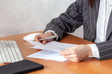 Closeup hands of woman in formal suit and white shirt, holding pen, sheets of paper, near computer keyboard, mouse and notebook. Concept of report writing, checking the documents, office job.の写真素材