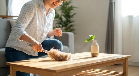 Smiling young woman actively cleaning coffee table with feather duster. Sunlight illuminates the dust particles. Woman maintains clean and tidy living room. Daily chores for a fresh home environment.の素材