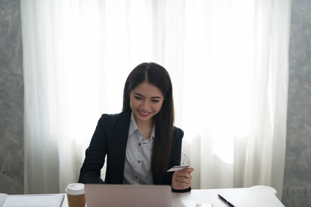 Happy beautiful asian young businesswoman holding credit card and using laptop computer in her workstation at desk. Online shopping conceptの写真素材