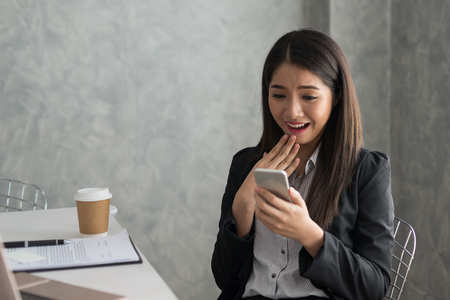 Excited asian business girl while reading a smart phone sitting in her workstation. Business Success Conceptの写真素材