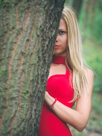 Blonde Woman with Red Dress in Oak Forest on a Summer Day Hiding Behind the Treeの写真素材