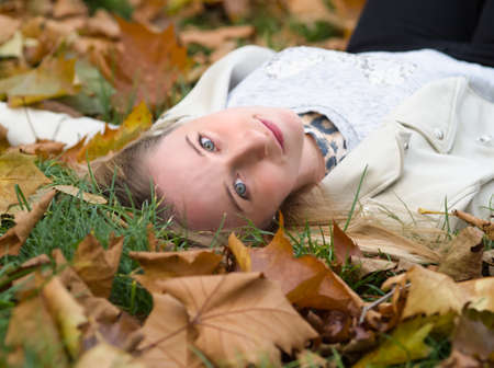Young Beautiful Blonde Woman Laying in the Grass in Autumn Park among Maple Leavesの写真素材