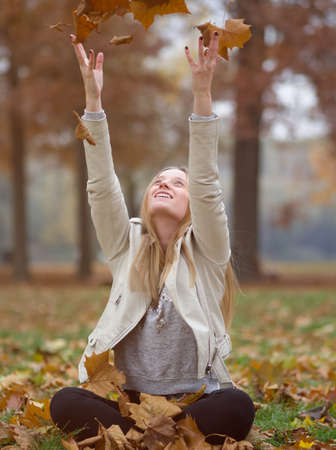 Young Blonde Woman Sitting in Autumn Park Throwing Brown Maple Leavesの写真素材