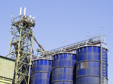 Large, blue industrial silos stand alongside a tall metal framework structure, symbolizing modern industry and storage under a clear blue sky.の写真素材