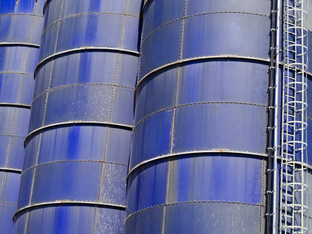 A detailed close-up shot of multiple blue industrial silos, highlighting their riveted metal panels and an industrial access ladder, under strong sunlight.の写真素材