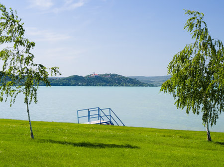 The picturesque free beach on Lake Balaton features a green lawn, elegant trees, and blue water stairs, with the iconic Tihany Abbey visible on the distant Tihany Peninsula under a clear summer sky.の写真素材