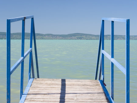 A close-up view of blue metal stairs and a wooden platform extending into the clear, turquoise waters of Lake Balaton, with distant hills visible under a bright blue sky.の写真素材