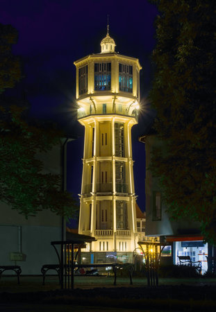 The illuminated SiÃ³fok Water Tower stands tall at night, framed by dark trees and surrounding buildings, with benches visible in the foreground under the deep blue sky.の写真素材