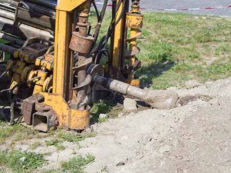 A heavy-duty yellow construction drill rig with an auger bit extends into the ground, surrounded by disturbed earth and green grass at a worksite under clear daylight.の写真素材