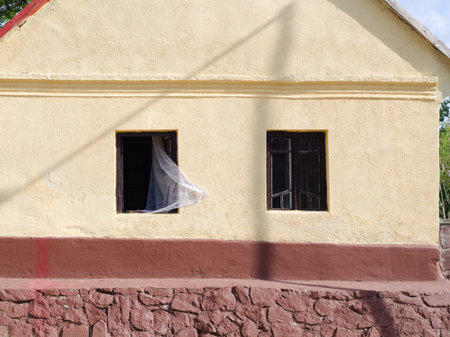 A weathered, light yellow building facade with a reddish-brown base and stone foundation, featuring two dark-framed windows, one with a white curtain softly blowing outwards.の写真素材