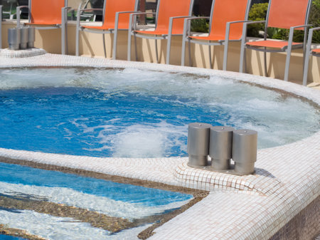 A close-up view of a resort bathtub with white and brown mosaic tiles, showcasing its inviting blue bubbling water and vibrant orange lounge chairs arranged in the blurred background under clear daylight.の写真素材