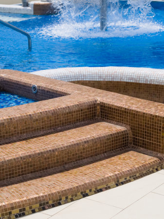 Elegant, brown mosaic-tiled steps lead into a bright blue swimming pool, with a modern stainless steel handrail and a splashing water feature visible in the background, creating an inviting scene.の写真素材