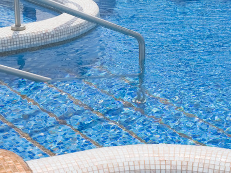 A close-up view of a vibrant blue swimming pool's mosaic-tiled steps and a gleaming metal handrail, framed by a white tiled edge in the foreground, indicating a clean and inviting aquatic area.の写真素材