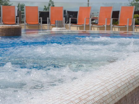 A close-up view of a bubbling hot tub with white and brown mosaic tiles, featuring blurred orange lounge chairs arranged in the background under clear daylight at a resort.の写真素材