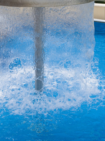 A dynamic close-up shot of a modern, metallic mushroom-shaped water feature, showing water vigorously cascading into a vibrant blue swimming pool below, highlighting the splashing motion.の写真素材
