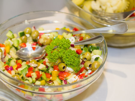 A vibrant close-up of a refreshing Mediterranean-style salad, generously filled with chopped vegetables, green olives, feta cheese, and garnished with fresh parsley in a clear glass bowl, ready for serving.の写真素材