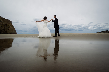 The bride and groom walk along the beach, reflecting in the water. Panoramic view of reflection. Walking along the beach against the rocks and ocean waves.の写真素材