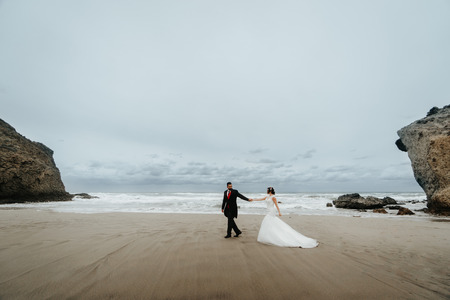 The bride and groom walk along the beach, reflecting in the water. Panoramic view of reflection. Walking along the beach against the rocks and ocean waves.の写真素材