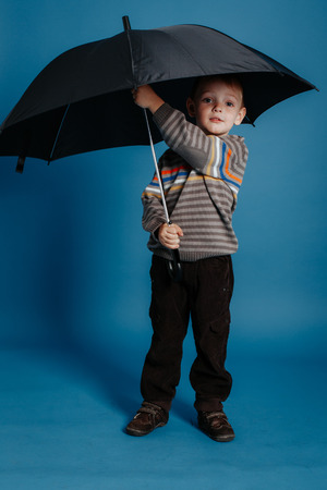 A little boy is standing under an open umbrella and smiling. On a blue backgroundの写真素材