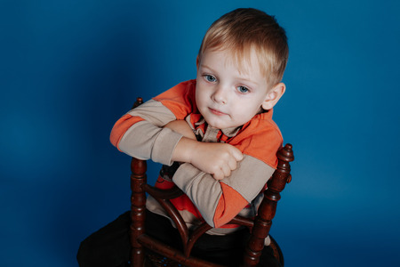 A little boy in a cap sits on a chair and thinks. On a blue backgroundの写真素材