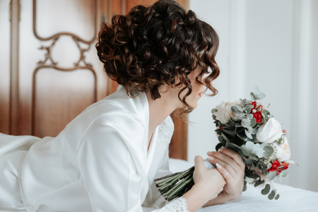 Bride with a bouquet of wedding flowers lying on the bed. Attractive young bride lies next to the dress and winter bouquet. Morning bride in a luxury hotelの写真素材
