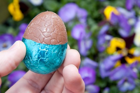 A chocolate easter egg held in the hand with colourful spring flowers in the background.の写真素材