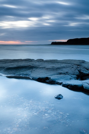 A generic costal scene with clouds, cliff, sea, some rocks and a pebble in a rock pool.  Taken after sunset so has a natural blue tint.の写真素材