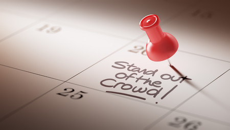 Concept image of a Calendar with a red push pin. Closeup shot of a thumbtack attached. The words Stand out of the crowd written on a white notebook to remind you an important appointment.の写真素材
