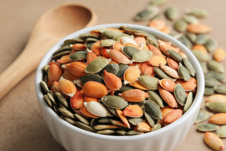 Roasted pumpkin seeds spilling from a bowl onto a table, next to a wooden spoon, creating a warm and inviting snack sceneの素材