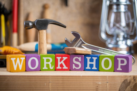 Hammer and wrench resting on colorful wooden toy blocks spelling the word workshop on workbenchの素材