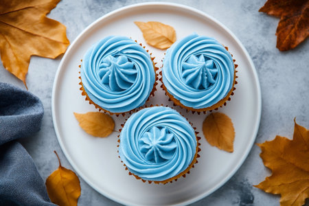Three cupcakes with blue icing are placed on a white plate, surrounded by autumn leaves, creating a sweet seasonal treatの素材