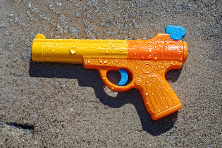 Colorful toy water gun left on the wet sand of a beach, creating a vibrant contrast against the textured surfaceの素材