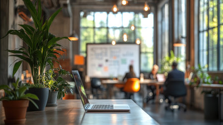 Open laptop computer sits on wooden desk with potted plants in a modern coworking office space with blurred business people meeting in backgroundの素材