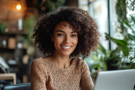 Young woman with curly hair smiling while working on a laptop in a modern cafe, enjoying the comfortable atmosphere and productive work sessionの素材