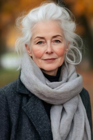 Portrait of an elegant senior woman wearing a gray scarf and a dark coat, enjoying a serene autumn day outdoorsの素材