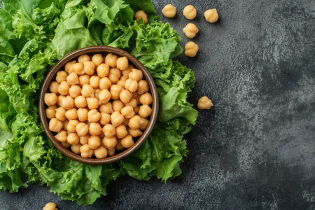 Healthy chickpeas in bowl resting on fresh green lettuce leaves on dark background, top view with copy spaceの素材