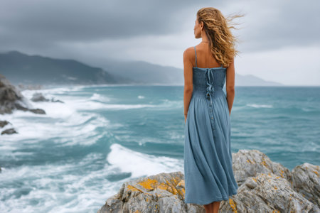 Woman stands on a rocky coast, gazing at the turbulent ocean waves crashing against the shore under a stormy skyの素材