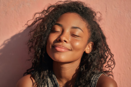Portrait of a young woman with curly hair, her eyes closed and a peaceful smile on her face, enjoying the warm sunlight against a pink wallの素材