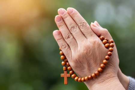 Hands holding a wooden rosary are praying in a natural setting with sunlightの素材