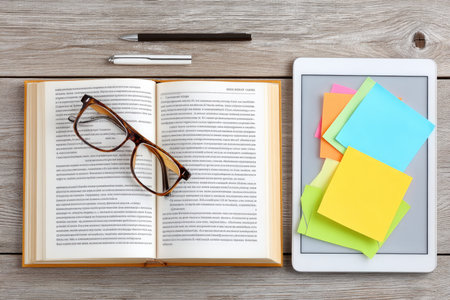 Office desk with open book, reading glasses, tablet and colorful sticky notes, studying and working conceptの素材