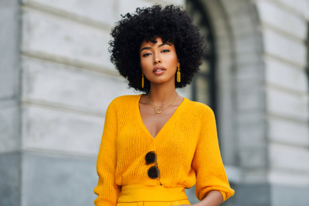 Fashionable businesswoman showcasing trendy yellow outfit and gold accessories in front of a modern buildingの素材
