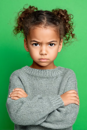 Young girl with curly hair expressing anger and defiance, standing with arms crossed on green backgroundの素材
