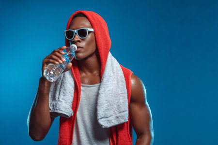 Sweaty fit man wearing sunglasses and a hoodie, hydrating with bottled water after exerciseの素材