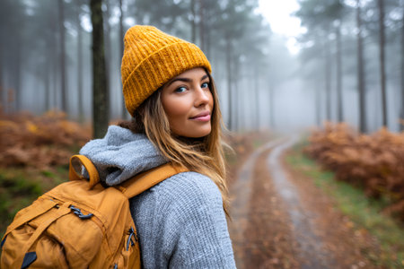 Woman with backpack traveling through foggy autumn forest trail, looking at cameraの素材