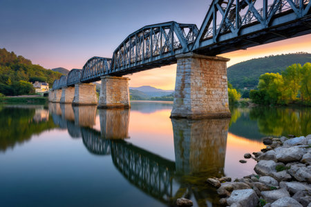 Metal truss bridge spanning a calm river with stunning reflections and warm sunset colorsの素材