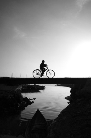 Silhouette of lonely young boy riding bicycle on small bridge cross the river at morning, he ride bike leisure,  this make sad, calm scene at countryside の写真素材