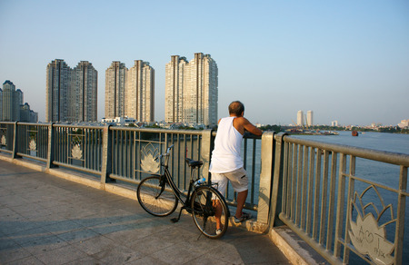 HO CHI MINH, VIET NAM- MAR 12  Healthy lifestyle of citizen at evening, ride bicycle to do exercise, stand on handrail of bridge to relaxed, enjoy a view, building at riverside, Vietnam, Mar 12, 2014のeditorial素材
