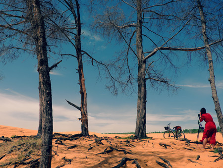 Woman traveling alone at Mui Ne, Vietnam, girl in red dress and selfie with bike on yellow sand hill under tree, tourist enjoy the trip in summetimeの写真素材
